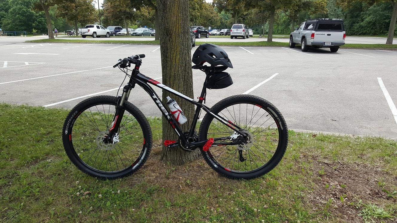 Trek X caliber 6: A black and red mountain bike leaning against a tree, with a helmet attached to the handlebars. A water bottle is mounted on the bike frame. In the background, a parking area with several cars is visible, surrounded by green grass and trees.