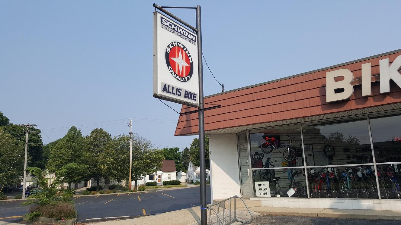 A storefront displaying the sign for "Schwinn Cycling and Fitness" and "Allis Bike," featuring a window with visible bicycles inside. The exterior has a brown shingle roof and there are trees in the background. The sky is clear with no clouds.