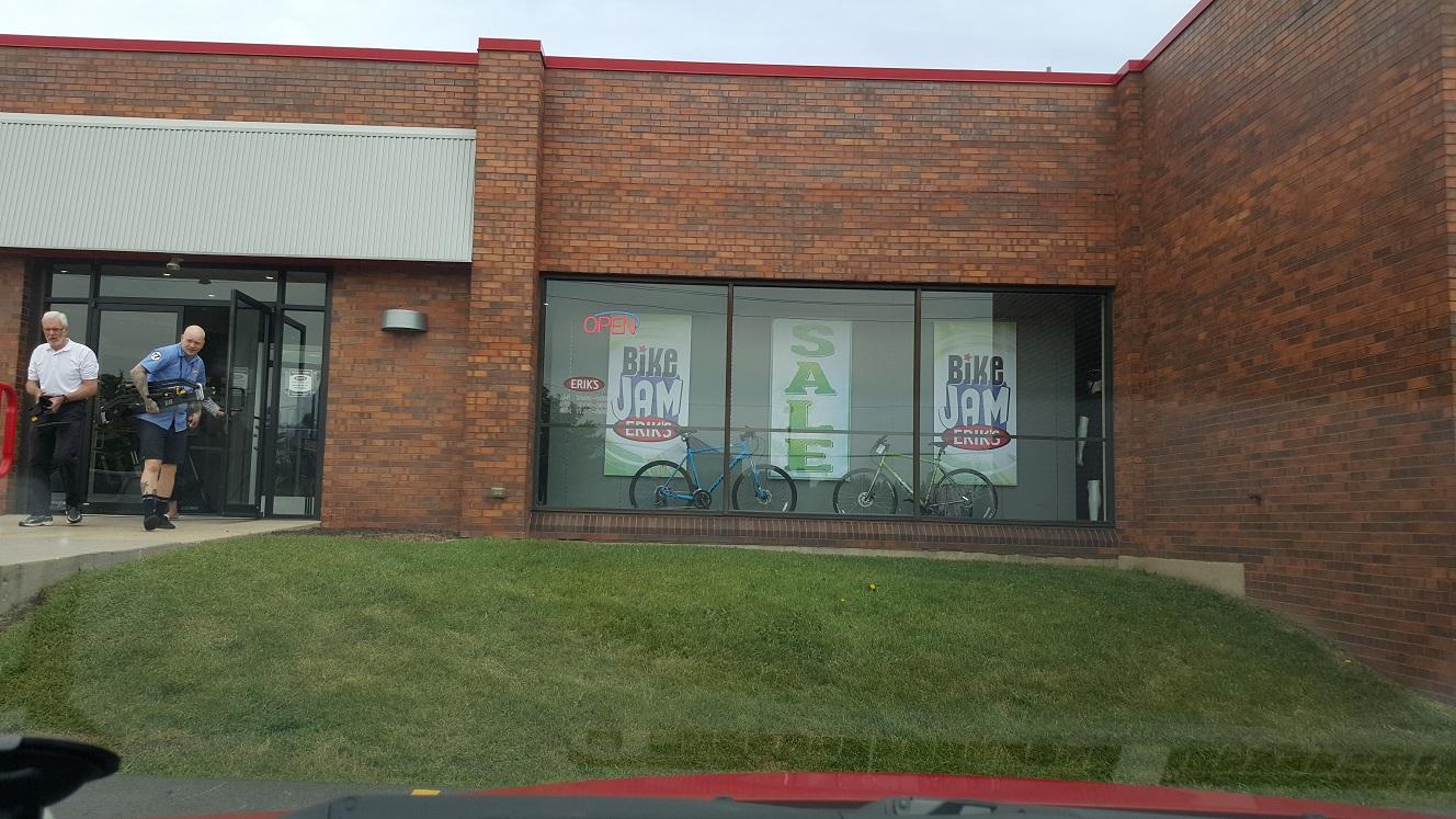 A storefront of a bicycle shop featuring a large "SALE" sign in the window. Two men are exiting the store, one carrying a bicycle. Visible bicycles in the display window are blue and green. The building is made of red brick, and the shop is identified as "Bike Jam." A grassy area is in front of the store.