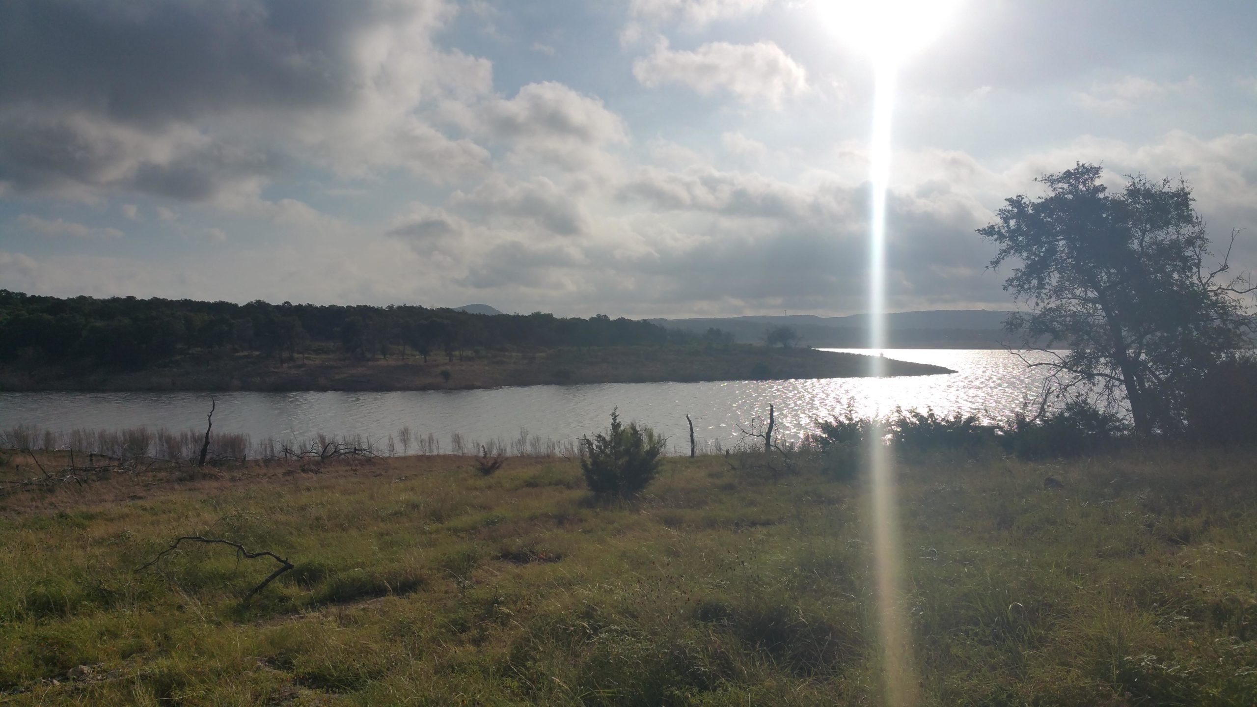 A scenic view of a calm lake surrounded by grassy land and sparse trees, under a cloudy sky with sunlight reflecting on the water. Madrone Trail mountain bike trail.
