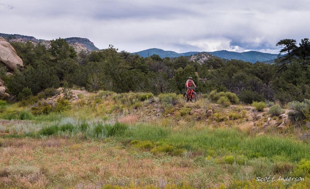 A cyclist on a red mountain bike rides along a rocky path through a lush, green landscape. The background features rolling hills under a cloudy sky, with dense trees dotting the scenery. Midland Hills Trails mountain bike trail.