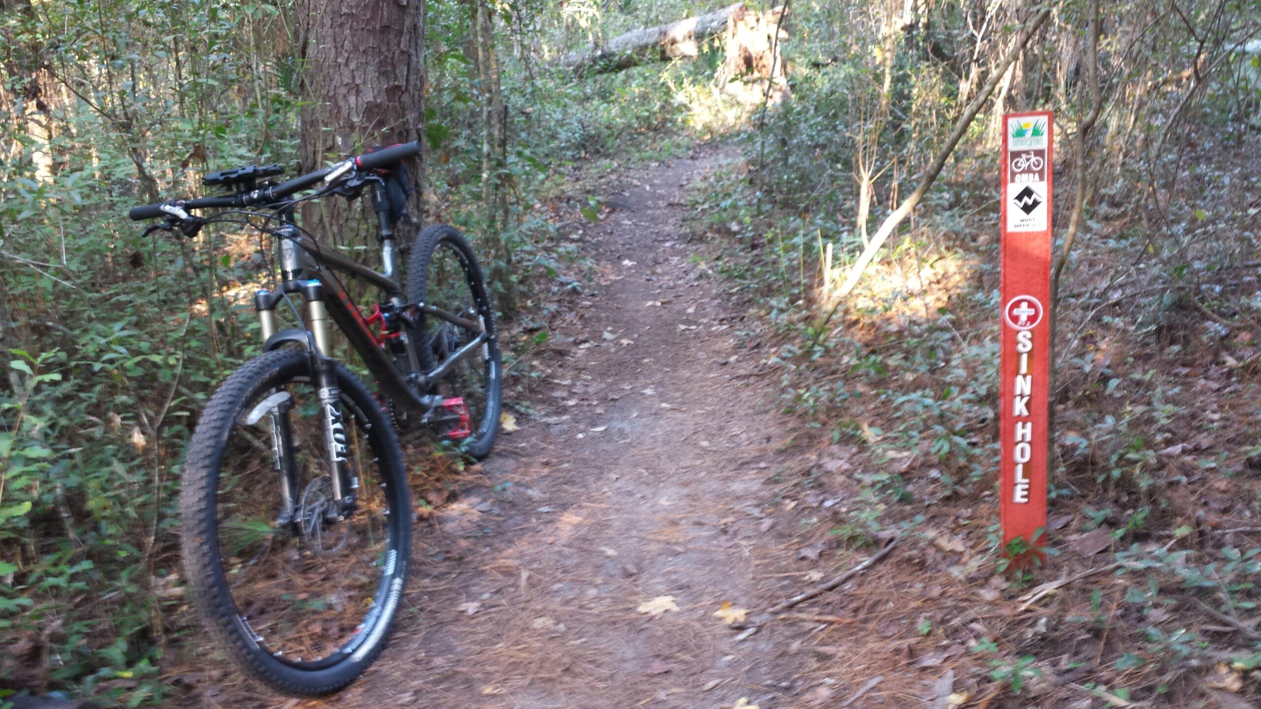 A mountain bike is parked next to a dirt trail surrounded by dense greenery. A trail marker labeled "SINKHOLE" is positioned on the right side, indicating caution ahead. The scene captures a serene outdoor environment ideal for biking activities. Santos mountain bike trail.