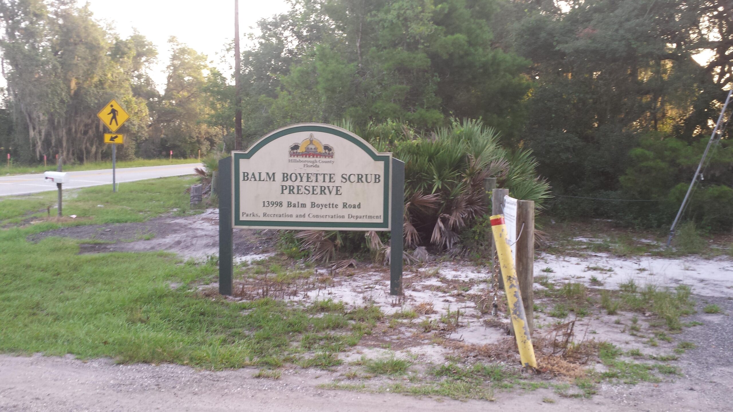 Sign for Balm Boyette Scrub Preserve located on Balm Boyette Road, featuring information about the preserve and a pedestrian crossing warning sign in the background. The area is surrounded by greenery and sandy ground, typical of a natural preserve setting. Balm Boyette Scrub Preserve mountain bike trail.