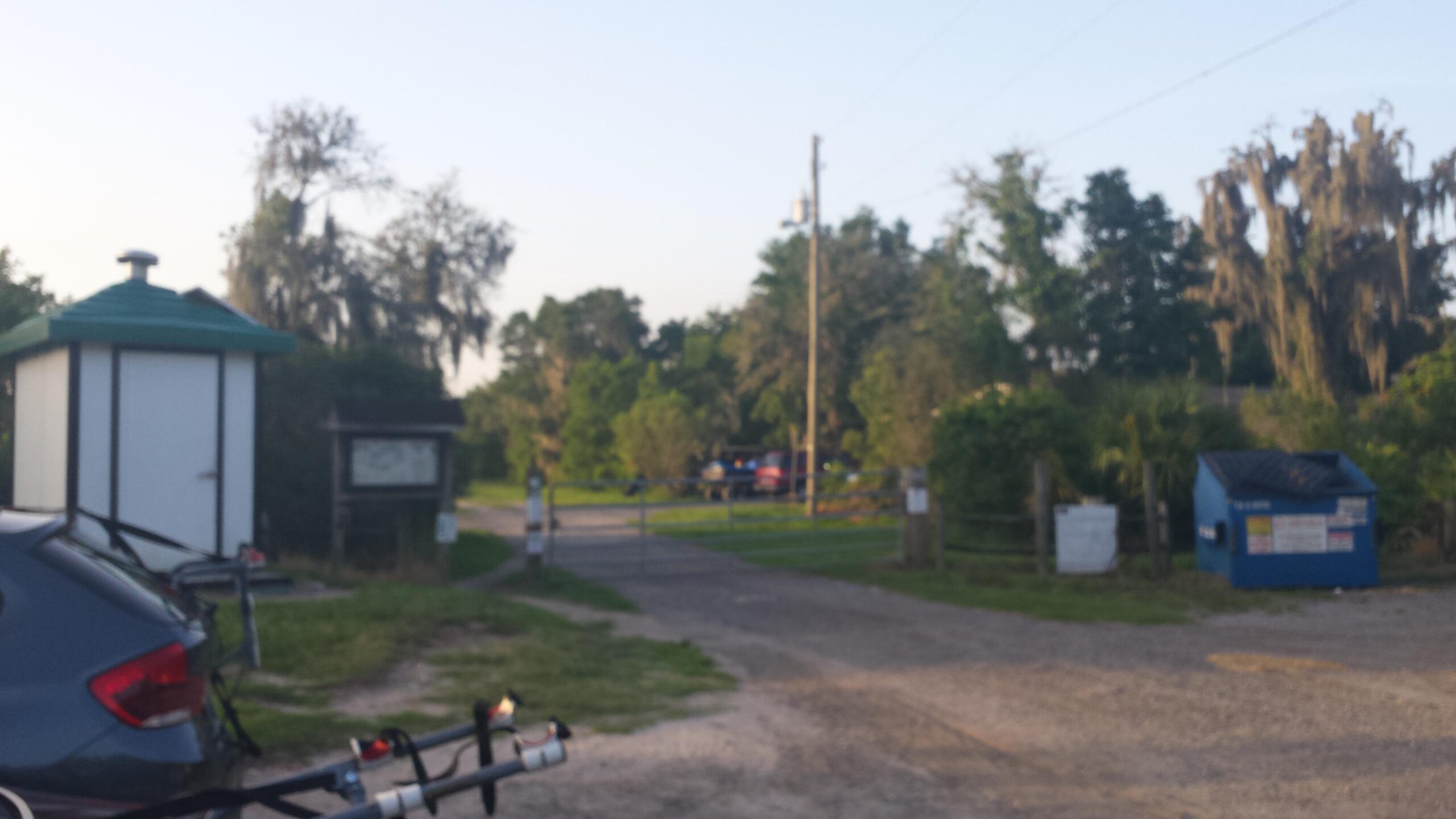 A view of a rural entrance with a gravel pathway leading to a closed gate, flanked by a small shelter and a blue dumpster. Lush greenery surrounds the area, with trees and bushes visible in the background. The scene suggests a quiet, natural environment in the early morning light. Balm Boyette Scrub Preserve mountain bike trail.