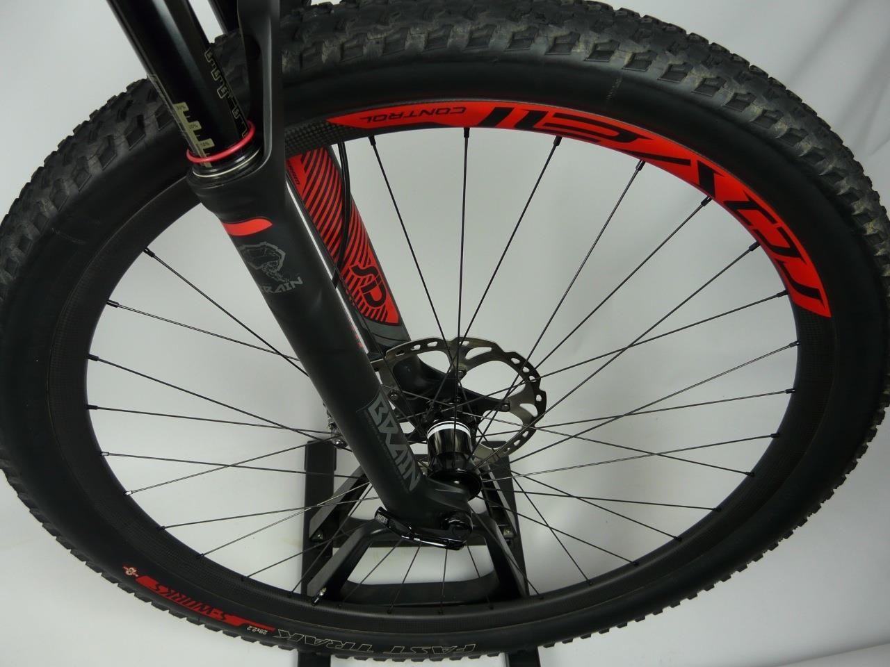Specialized Epic Expert Carbon 29er: Close-up view of a mountain bike front wheel, featuring a black fork and red accents. The tire has a rugged tread pattern, suitable for off-road terrain, and the rim is marked with the brand name. The background is plain, highlighting the details of the bike components.