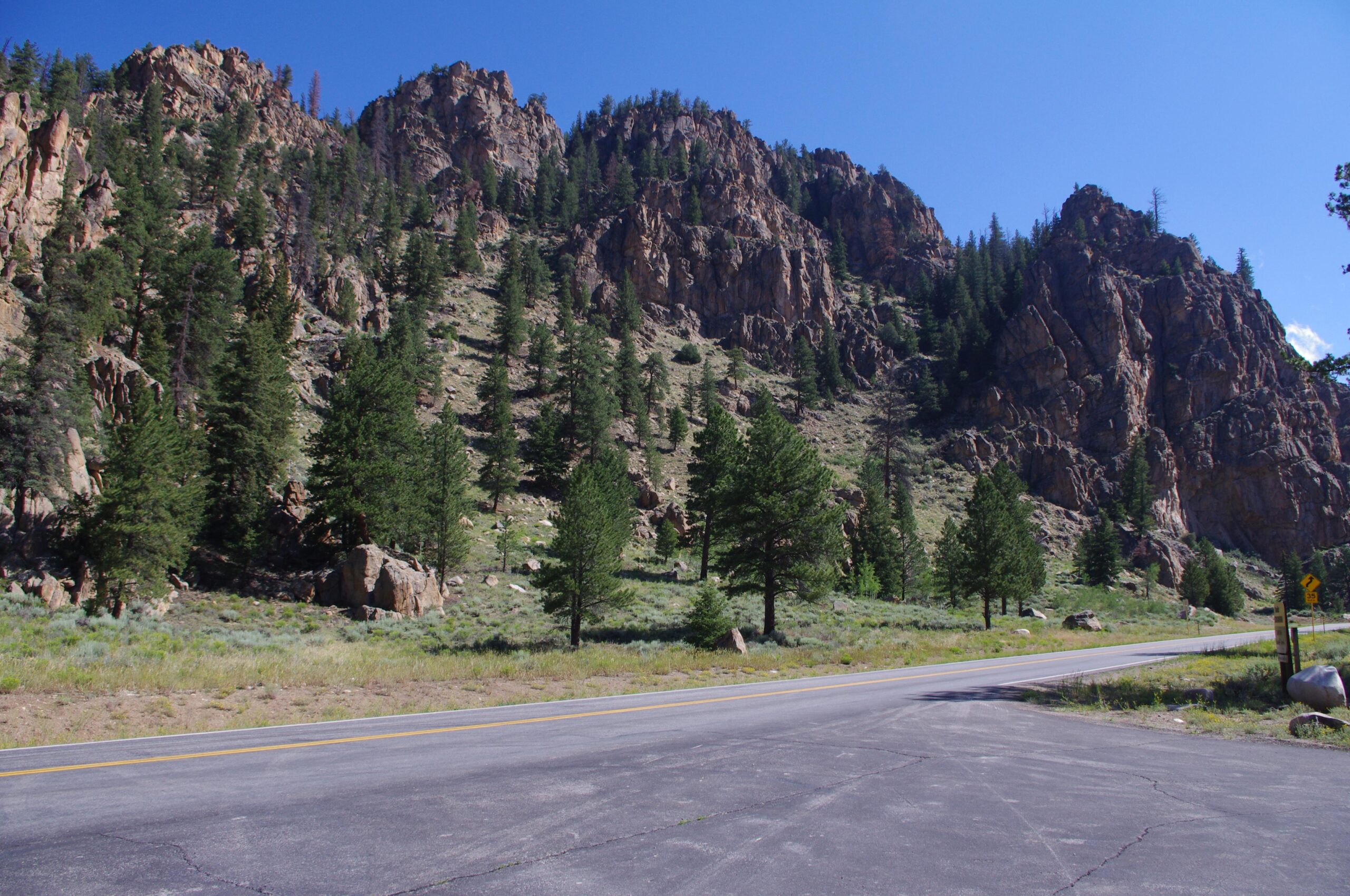 A scenic view of a mountainous landscape featuring rocky cliffs and dense coniferous trees under a clear blue sky, with a portion of a paved road in the foreground. Doctor Park mountain bike trail.
