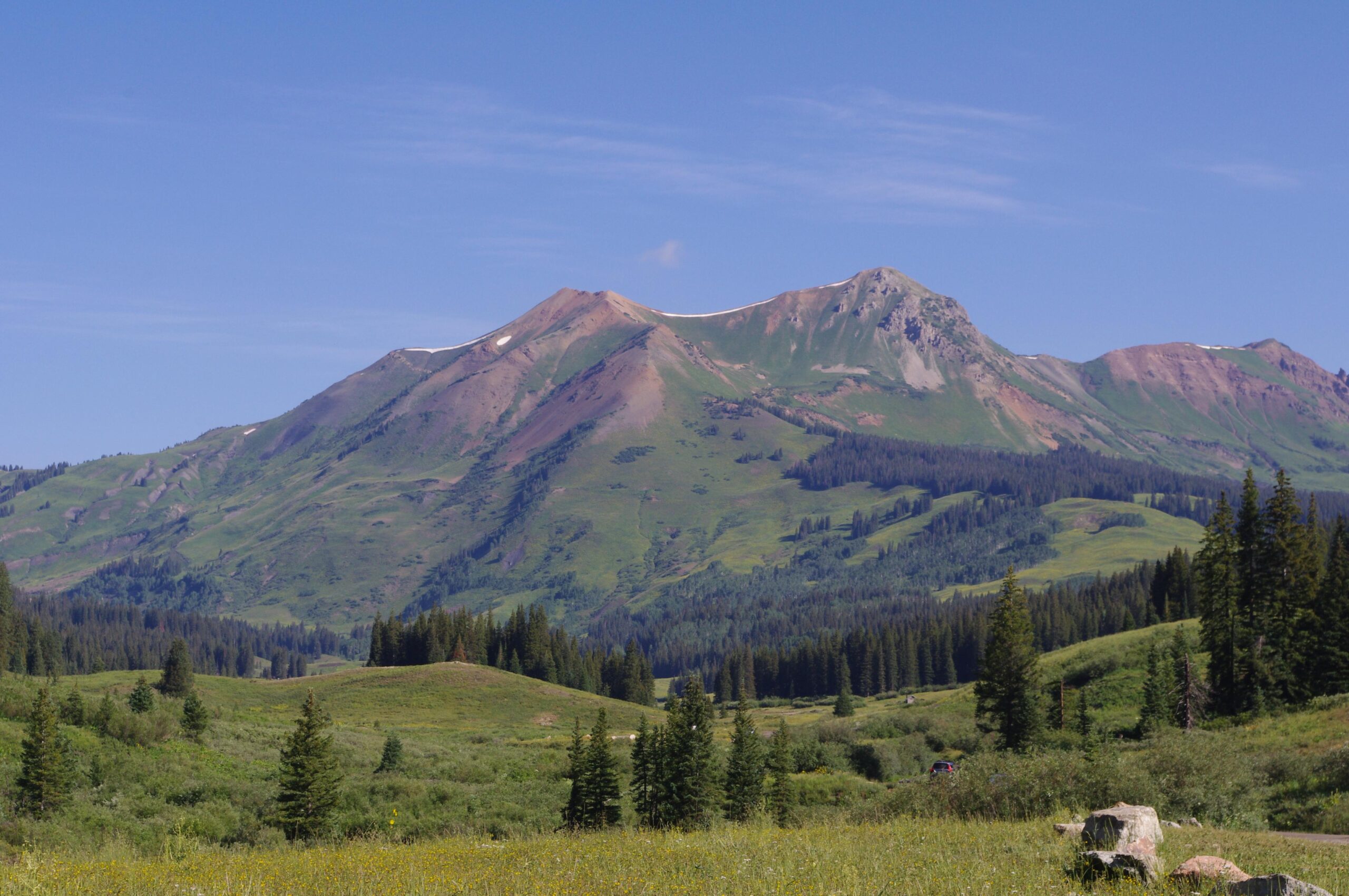 A scenic view of a mountainous landscape featuring grassy slopes, rugged peaks, and patches of snow under a clear blue sky. The foreground includes lush greenery and evergreen trees, leading to the base of the mountains. Trail 401 mountain bike trail.