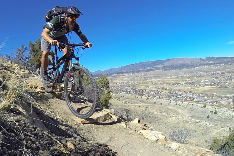 A mountain biker navigating a rocky trail with a scenic view of a valley and mountains in the background. The cyclist, wearing a helmet and sunglasses, is focused and in motion, with a backpack secured on his back. The sky is clear and blue, highlighting the outdoor adventure setting.