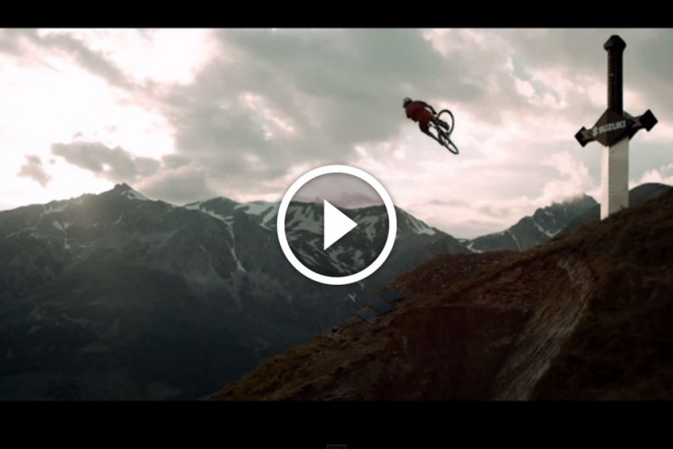 A mountain biker performs a jump over a rocky terrain, with dramatic mountain scenery in the background and a cross-shaped monument on the right. The sky is partly cloudy, adding to the adventurous atmosphere of the scene.