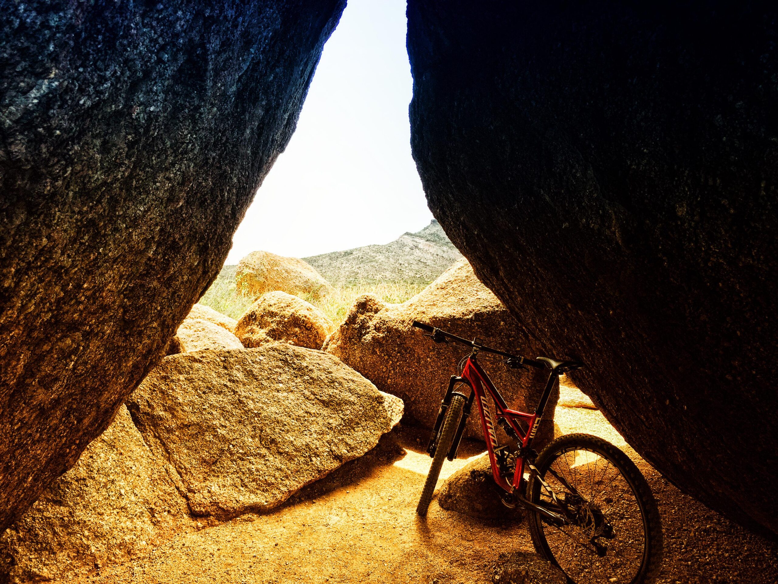 A mountain bike resting on sandy ground between large, rugged boulders, with a bright sky visible in the background. The scene captures the natural terrain and rocky landscape. Brown's Ranch to Granite Mountain mountain bike trail.