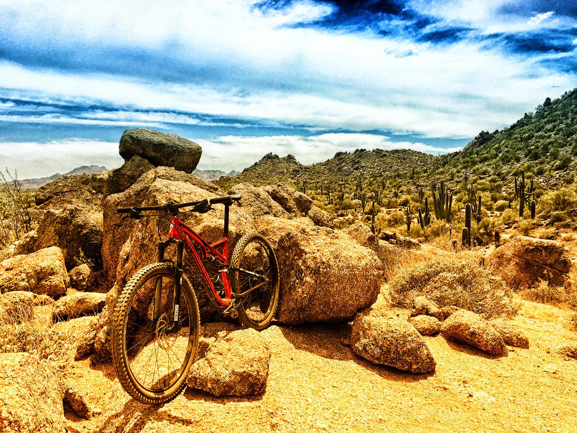 A red mountain bike resting against large rocks in a desert landscape, with cacti and rugged mountains in the background under a partly cloudy sky. Brown's Ranch to Granite Mountain mountain bike trail.