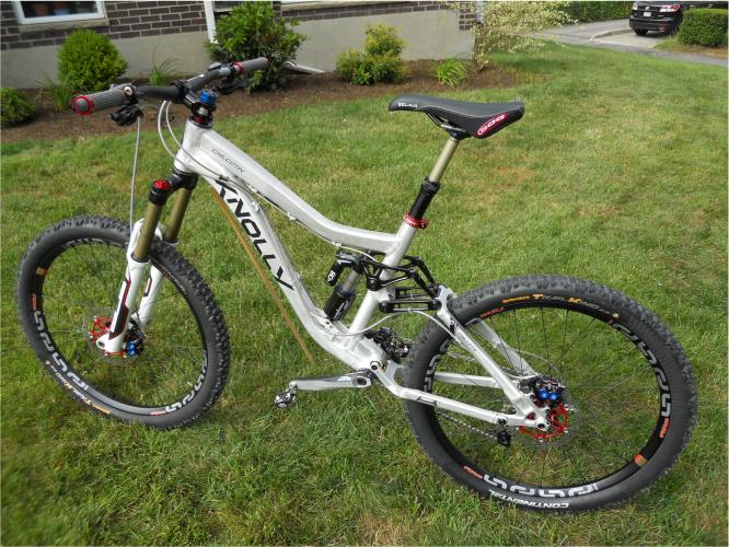 Knolly Chilcotin: A mountain bike with a silver frame, black and red handlebars, and thick tires, positioned on a grassy area. The bike features a suspension system and is parked near a residential home with greenery in the background.