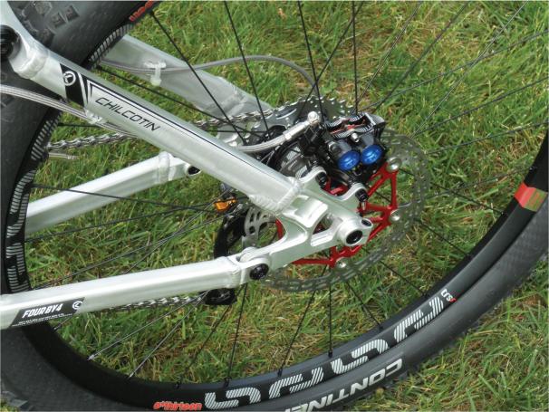 Knolly Chilcotin: Close-up view of the rear wheel of a mountain bike, showcasing the silver frame, disc brake, and tire. The bike features detailed components including a red brake caliper and an intricate arrangement of gears and cables, set against a green grass background.