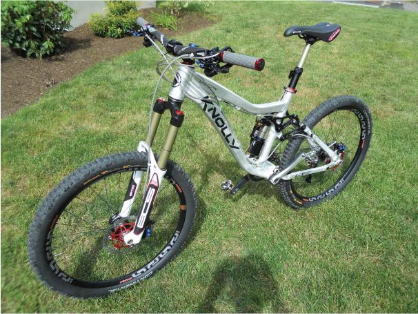 Knolly Chilcotin: A silver mountain bike with prominent suspension, rugged tires, and a sleek frame is positioned on a grassy area. The bike features red and black accents, including the handlebars and seat. Lush greenery surrounds the scene.