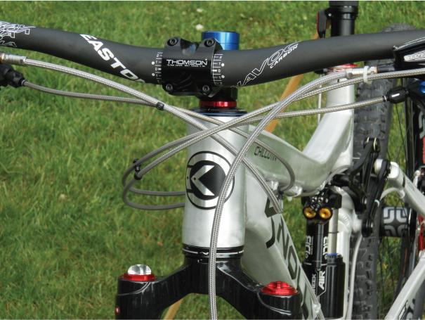 Knolly Chilcotin: Close-up view of a bicycle's handlebars and stem, featuring a silver frame with visible components including brake cables and a blue top cap. The handlebar is branded with “Easton,” and the stem has “Thomson” visible, showcasing a well-maintained mountain bike resting on green grass.