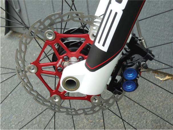 Knolly Chilcotin: Close-up image of a bicycle's front brake system, featuring a red disc brake rotor attached to a black and white fork. The rotor is designed with a star pattern, and the brake caliper includes blue hydraulic pistons. The background shows a textured surface.