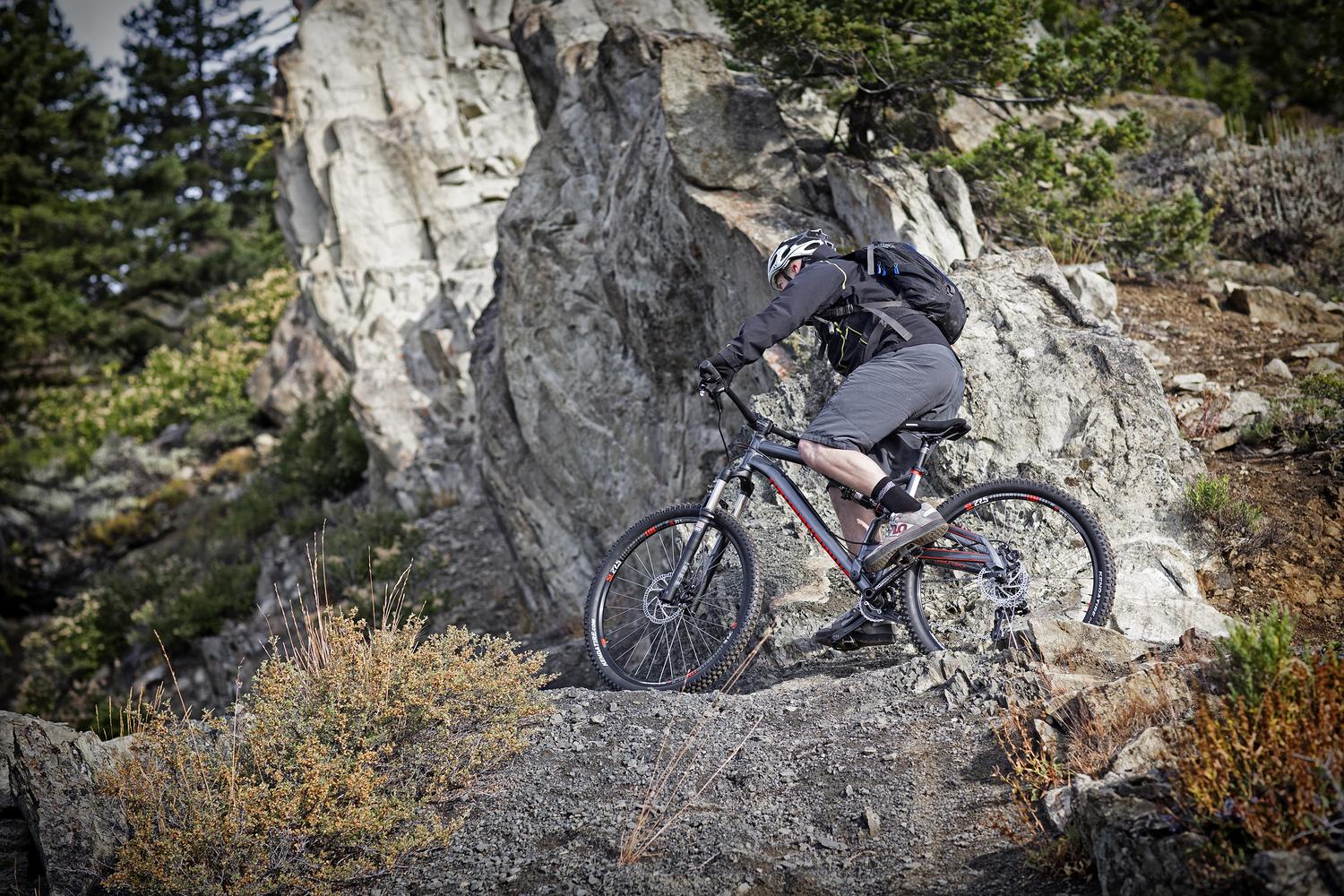 Diamondback Atroz Comp: A mountain biker navigating a rocky terrain, wearing a helmet and a backpack. The background features a mix of rocky outcrops and greenery, illustrating an outdoor trail setting.