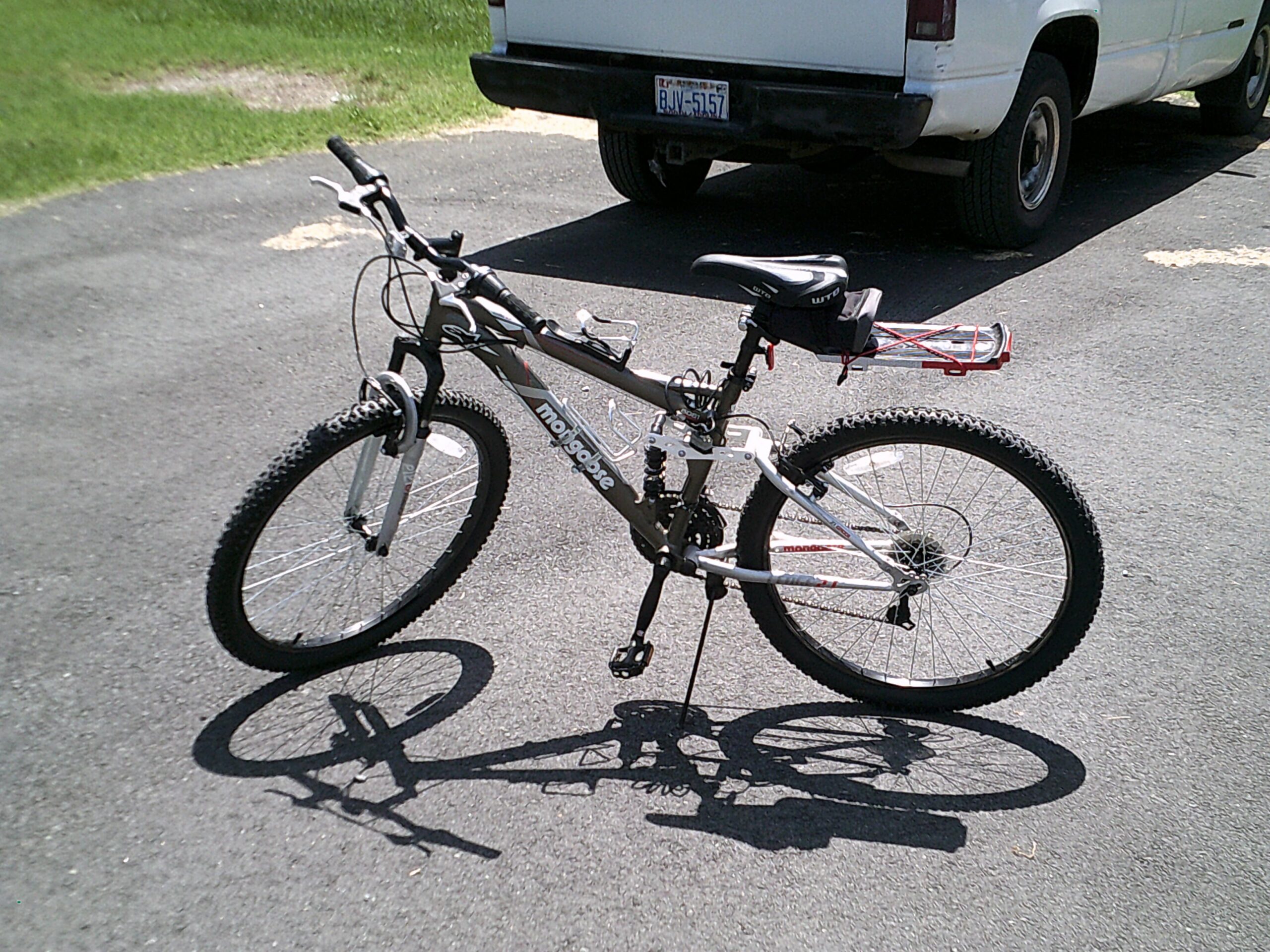 Mongoose Mongoose Ledge 2.1: A mountain bike parked on a paved surface, with its shadow visible. The bike features a grey frame, thick tires, and a rear rack with a small bag attached. In the background, a white vehicle is partially visible, along with green grass. The scene is well-lit, indicating a sunny day.