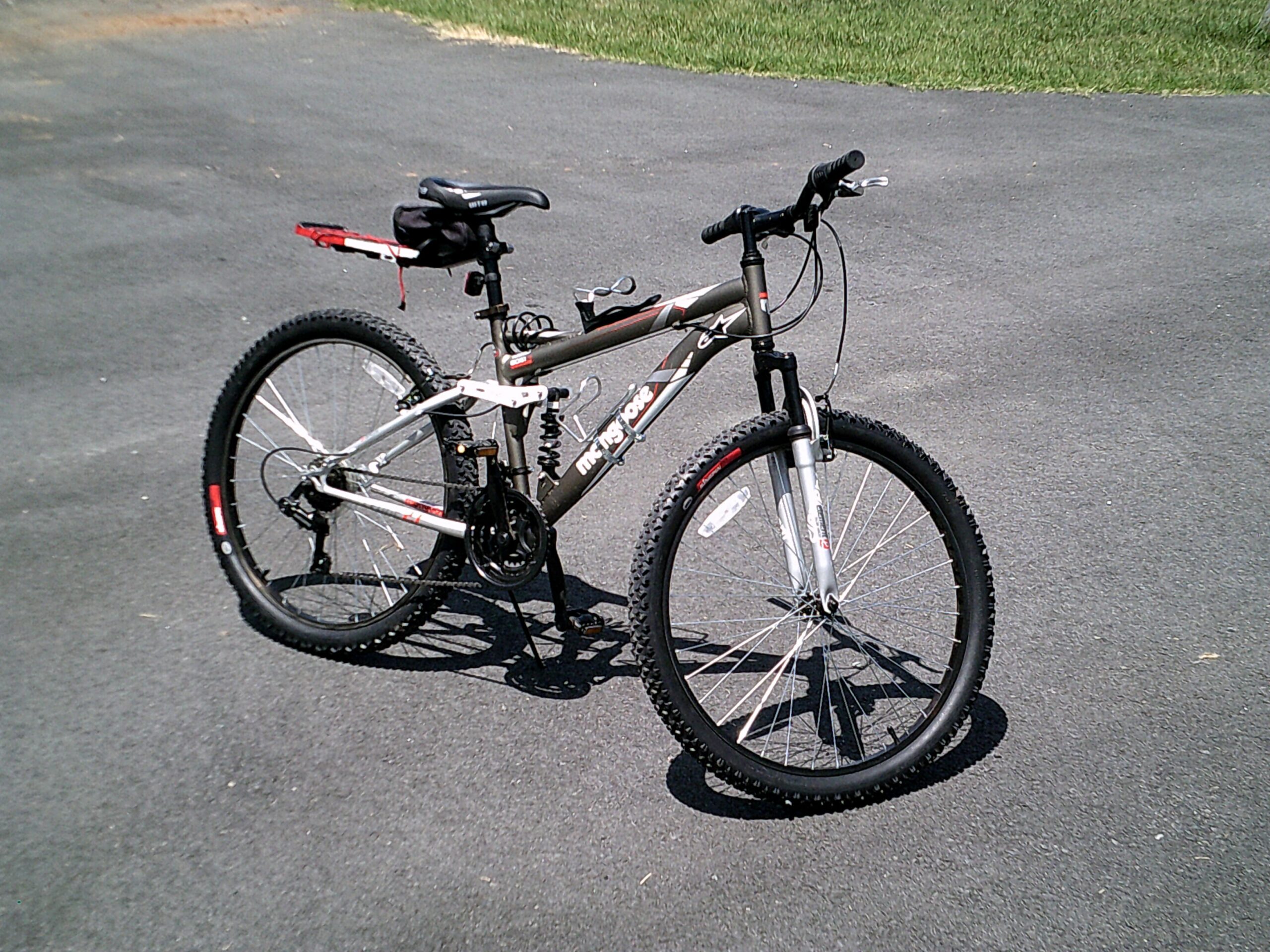 Mongoose Mongoose Ledge 2.1: A mountain bike parked on an asphalt surface, featuring a grey frame, black tires, and a rear rack with a small bag. The background shows a patch of grass.