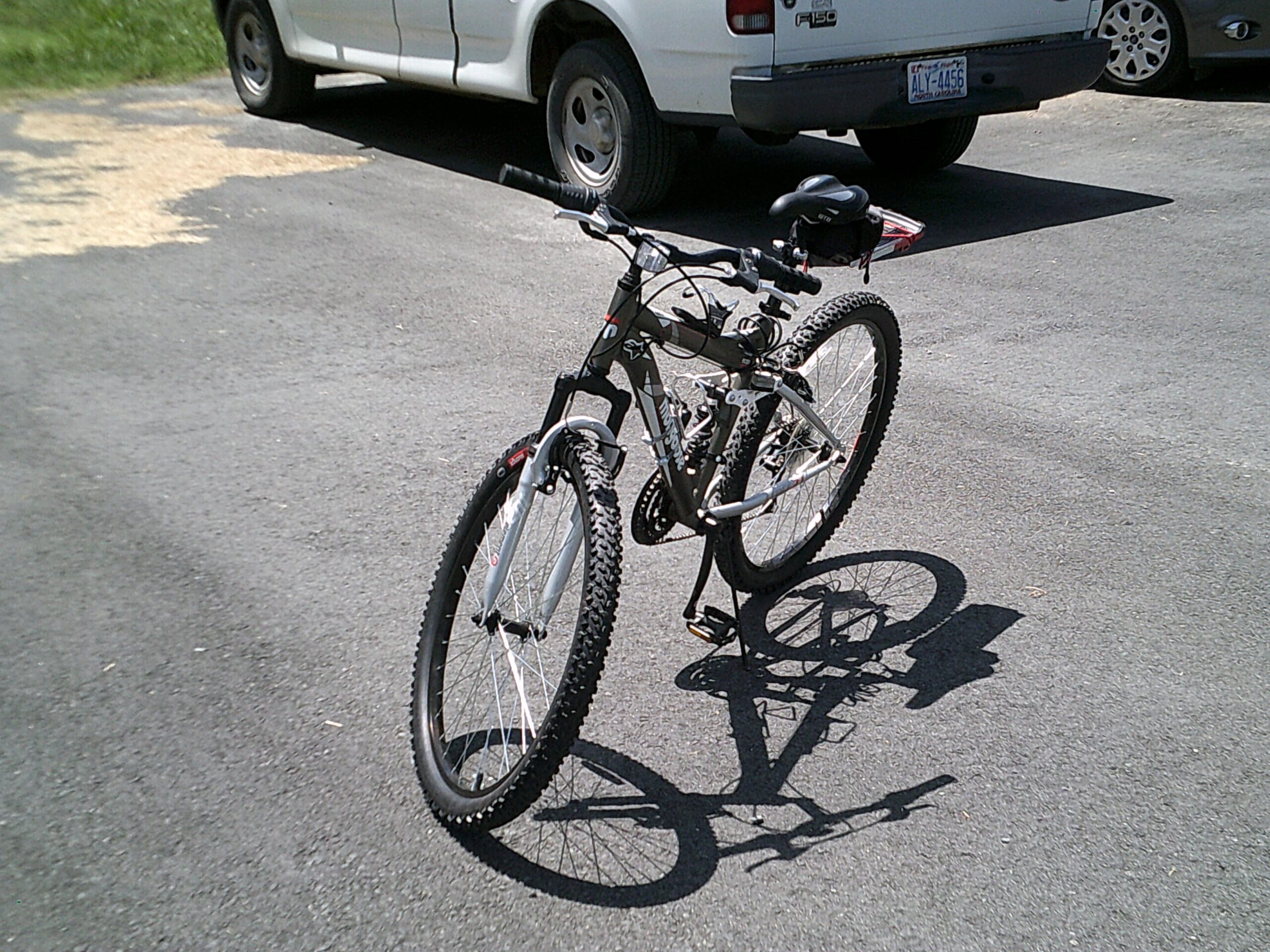 Mongoose Mongoose Ledge 2.1: A black mountain bike parked on a paved surface, with a white pickup truck and a gray car in the background. The bike has thick tires and is positioned upright, casting a shadow on the ground.