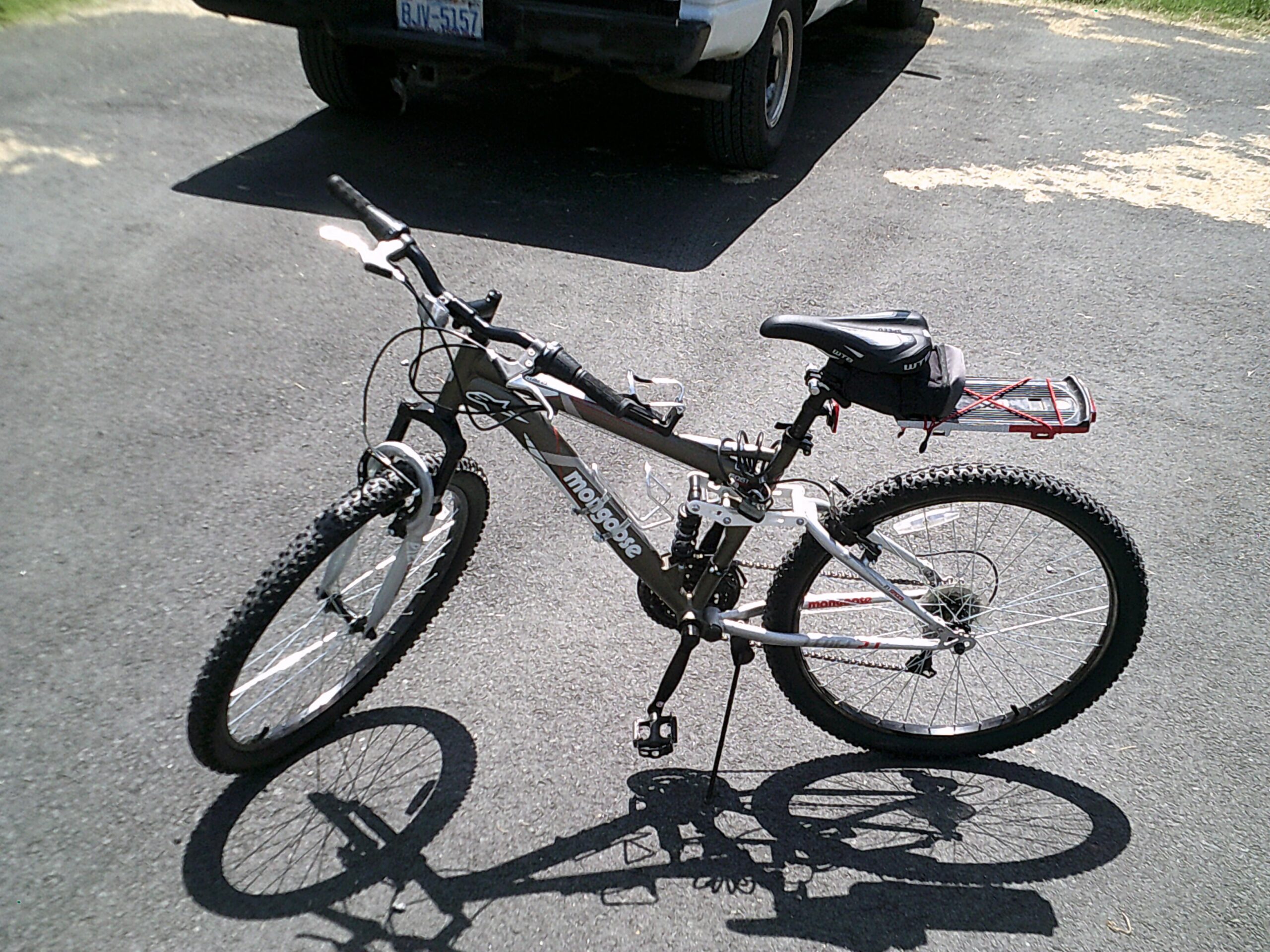 Mongoose Mongoose Ledge 2.1: A gray mountain bicycle with black and white accents, featuring thick tires, a comfortable seat, and a small storage bag on the rear. The bike is positioned on a black asphalt surface, with a pickup truck partially visible in the background.