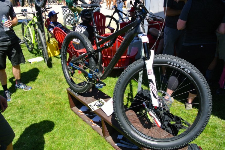 A black mountain bike displayed on a wooden stand, showcasing its front wheel and suspension fork. The background features people and other bicycles in a sunny outdoor setting, with green grass and red chairs visible.