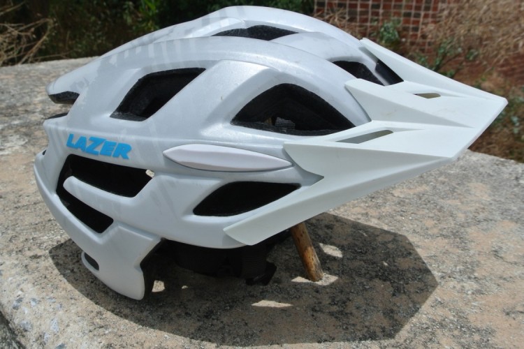 A white cycling helmet with ventilation openings and a sleek design, displayed on a stone surface. The helmet features the brand name "LAZER" prominently in blue. The background shows natural greenery and hints of a rustic setting.