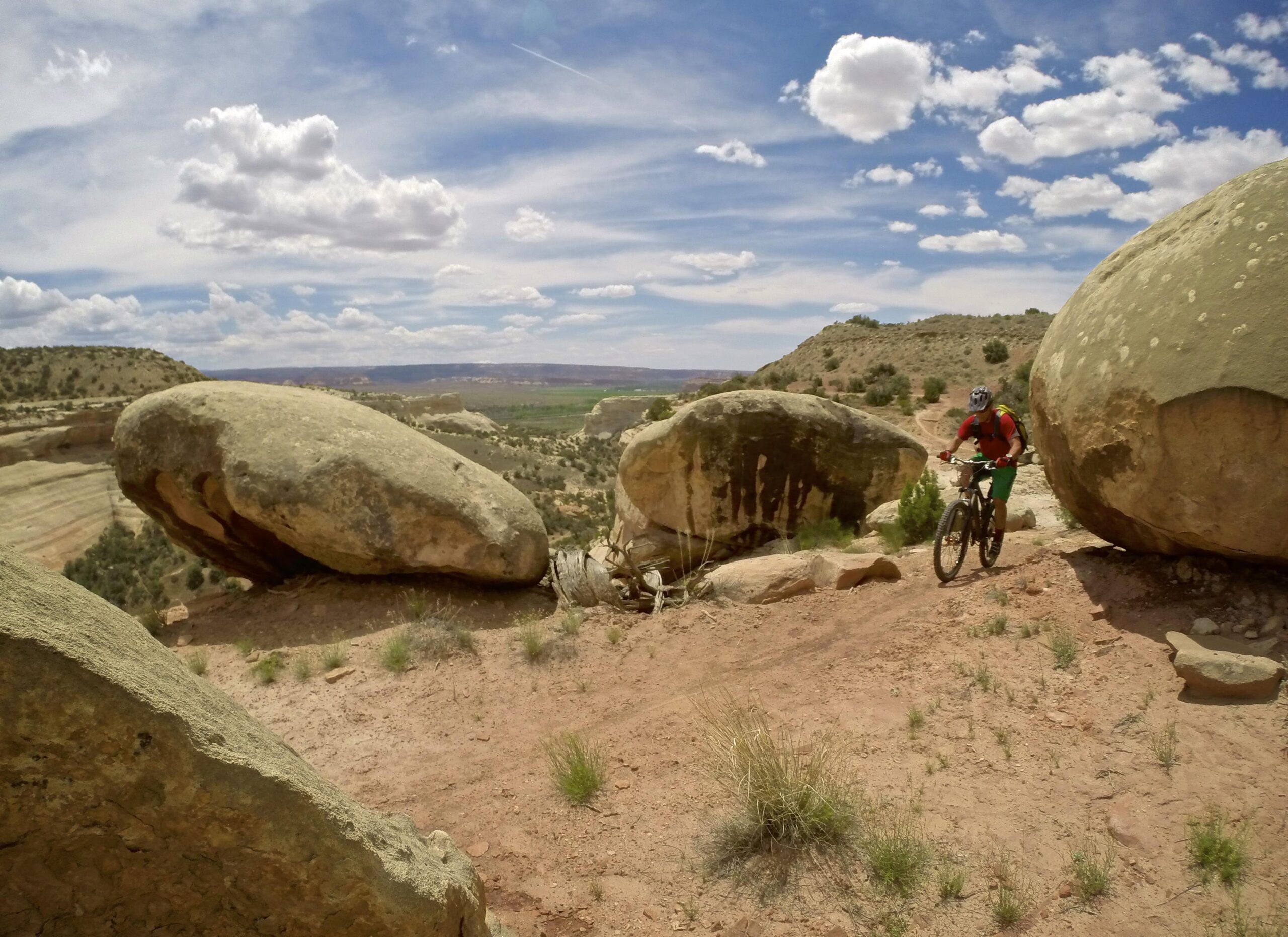 A mountain biker riding on a dirt trail surrounded by large boulders, with a scenic view of hills and a partly cloudy sky in the background. Western Rim mountain bike trail.