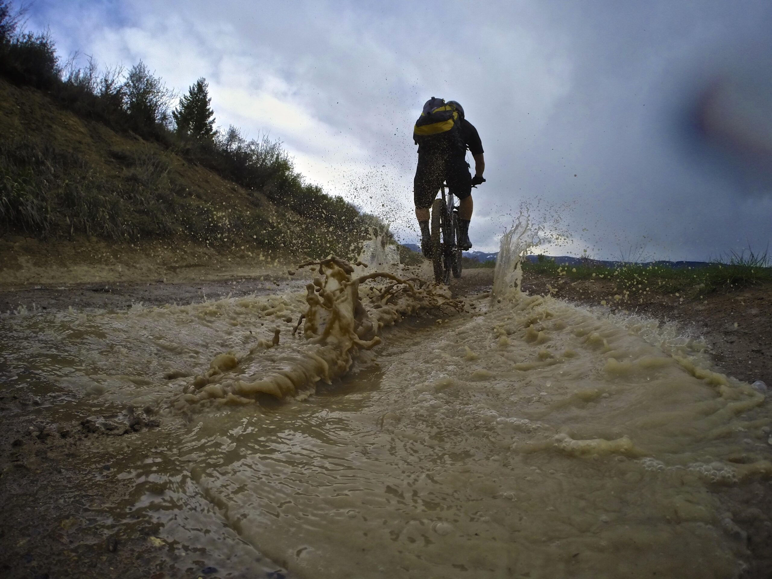 A cyclist riding through a muddy trail, splashing water and mud up from the ground, with a cloudy sky in the background. The image captures the dynamic action of mountain biking in a natural environment. Berry Creek Loop mountain bike trail.