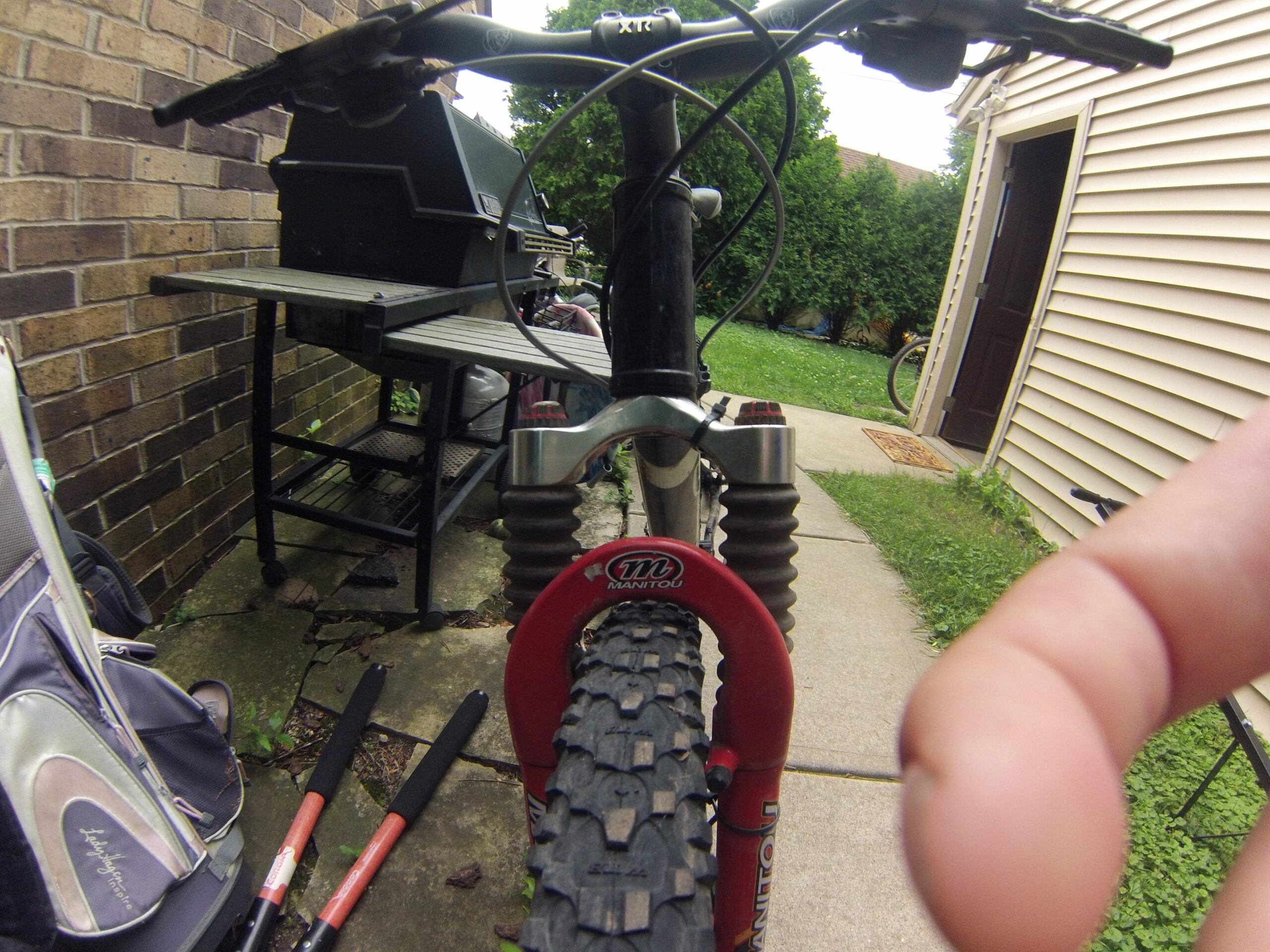 Photo of a mountain bike viewed from the front, featuring a close-up of the front tire and suspension. In the background, a black barbecue grill is visible next to a house, with some greenery and a visible pathway. A finger is partially in the frame, suggesting a point of focus.
