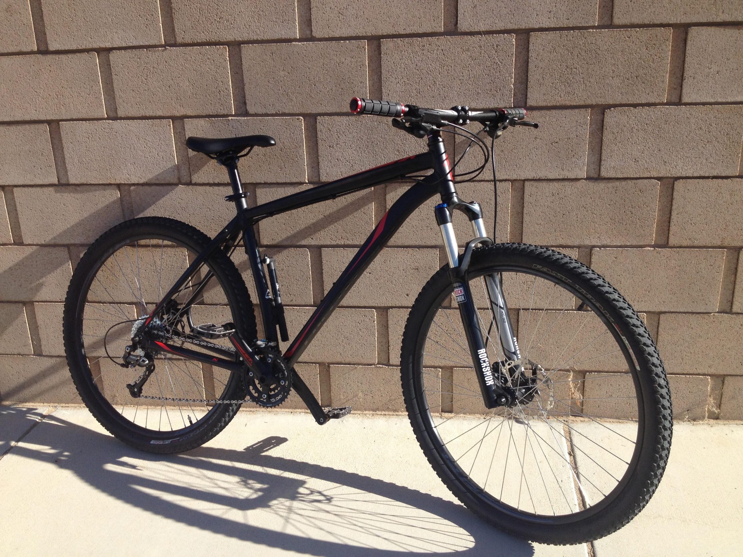 Specialized Hardrock Sport Disc 29er: A black mountain bike with red accents, leaning against a textured beige wall. The bike features thick, knobby tires and a front suspension fork, with a single gear setup visible on the rear wheel. The sunlight casts a shadow of the bike onto the concrete ground.