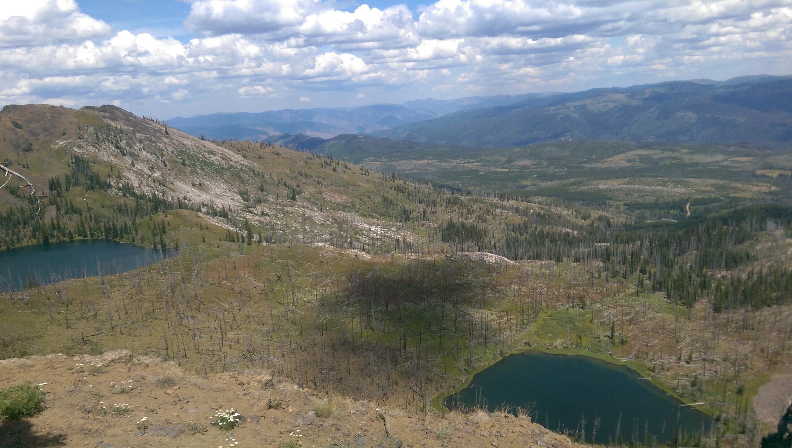A panoramic view of a mountainous landscape featuring two blue lakes surrounded by patches of green and dry land. The foreground shows rocky terrain with sparse vegetation, while the background features rolling hills and a vast sky filled with scattered clouds. Some areas appear to have been affected by past wildfires, with several dead trees visible. Lava Ridge Trail #149 mountain bike trail.