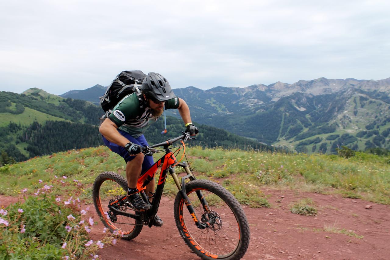 A mountain biker descends a rocky trail surrounded by lush green hills and distant mountains, wearing a helmet and backpack. Wildflowers bloom along the path as the cyclist leans forward to navigate the terrain. Wasatch Crest mountain bike trail.