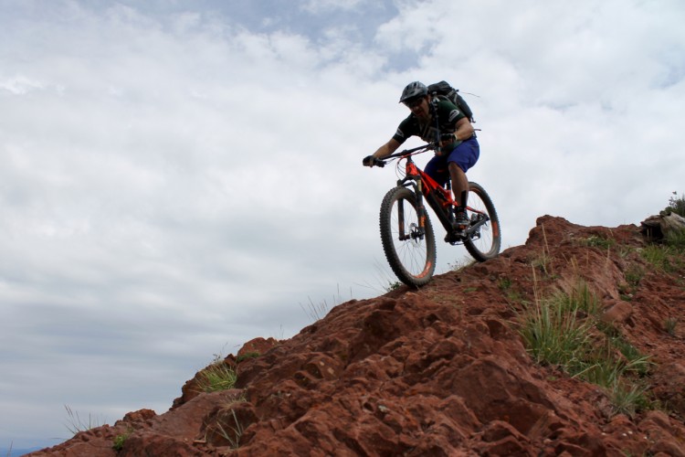 A mountain biker riding down a rocky terrain and navigating steep inclines, with a cloudy sky in the background. The rider is wearing a helmet and backpack, showcasing an adventurous outdoor activity.