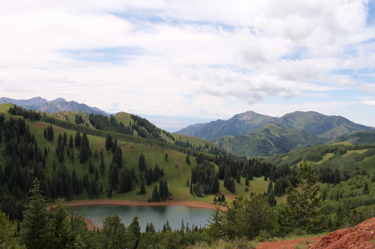 A scenic view of rolling green hills and mountains under a partly cloudy sky, featuring a serene lake surrounded by evergreen trees. Wasatch Crest mountain bike trail.