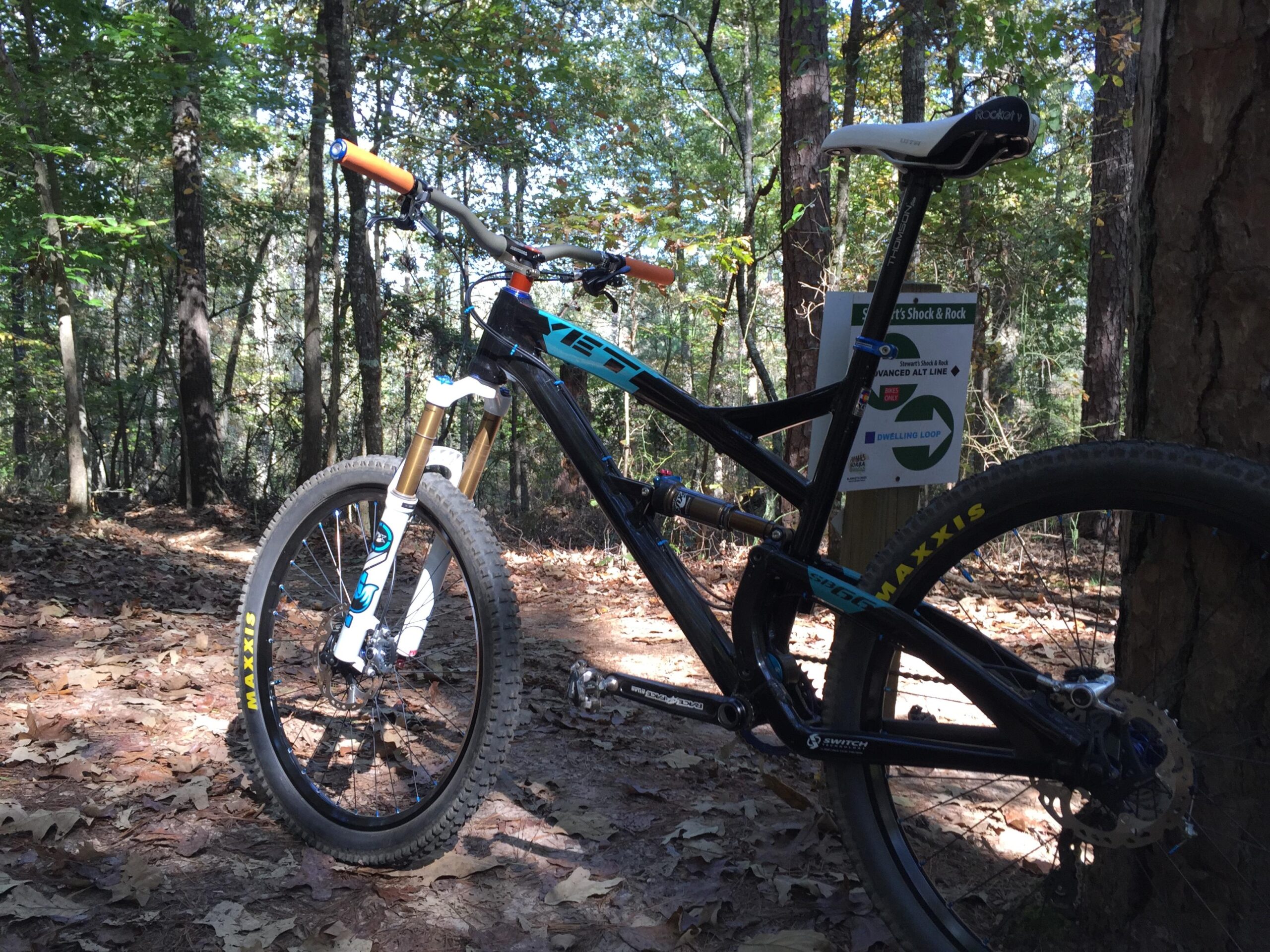 Yeti SB66: A mountain bike parked on a leaf-covered trail in a wooded area, with trees in the background. A trail sign is visible in the background, indicating an advanced line and a dwelling loop. The bike features a black frame with blue and orange accents, and fat tires labeled 'Maxxis'.