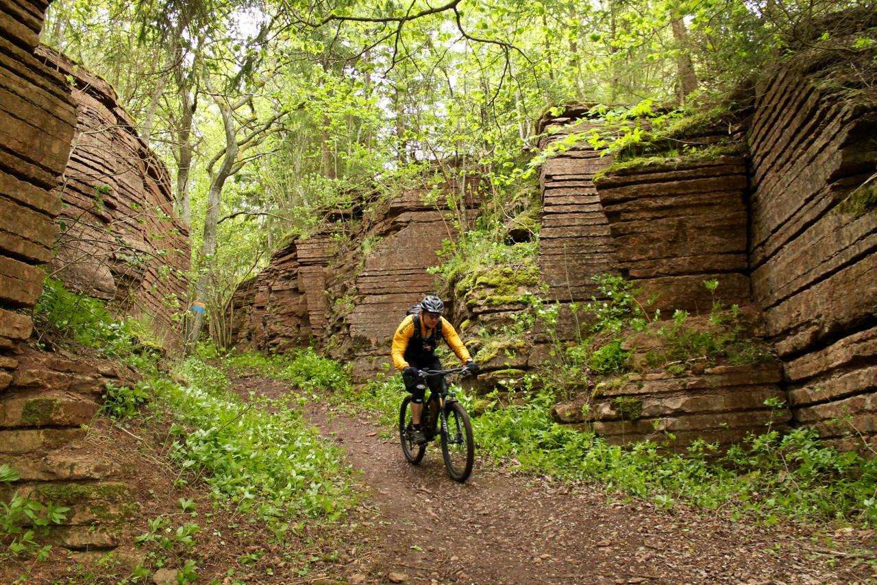 A mountain biker rides along a narrow, rocky trail flanked by tall, layered stone formations and lush greenery in a forested area. Light filters through the trees, creating a vibrant, natural atmosphere. Kinnekulle Trail System mountain bike trail.