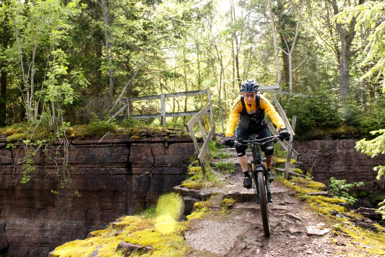 A mountain biker crossing a narrow, moss-covered path next to a steep rock face, surrounded by lush greenery and trees. A wooden bridge is visible in the background, highlighting the natural setting. Kinnekulle Trail System mountain bike trail.