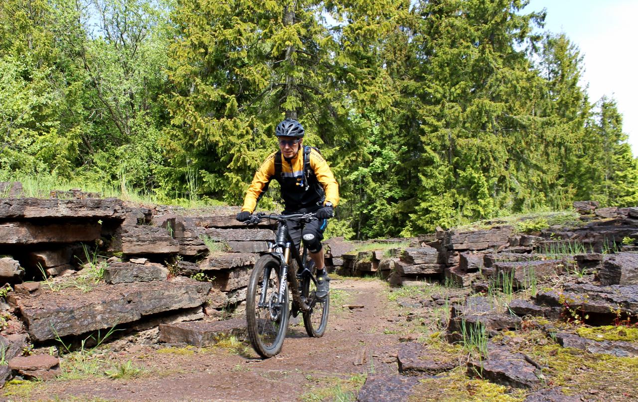 A mountain biker in yellow and black gear rides along a rocky trail in a forested area, surrounded by green trees and moss-covered rocks. Kinnekulle Trail System mountain bike trail.