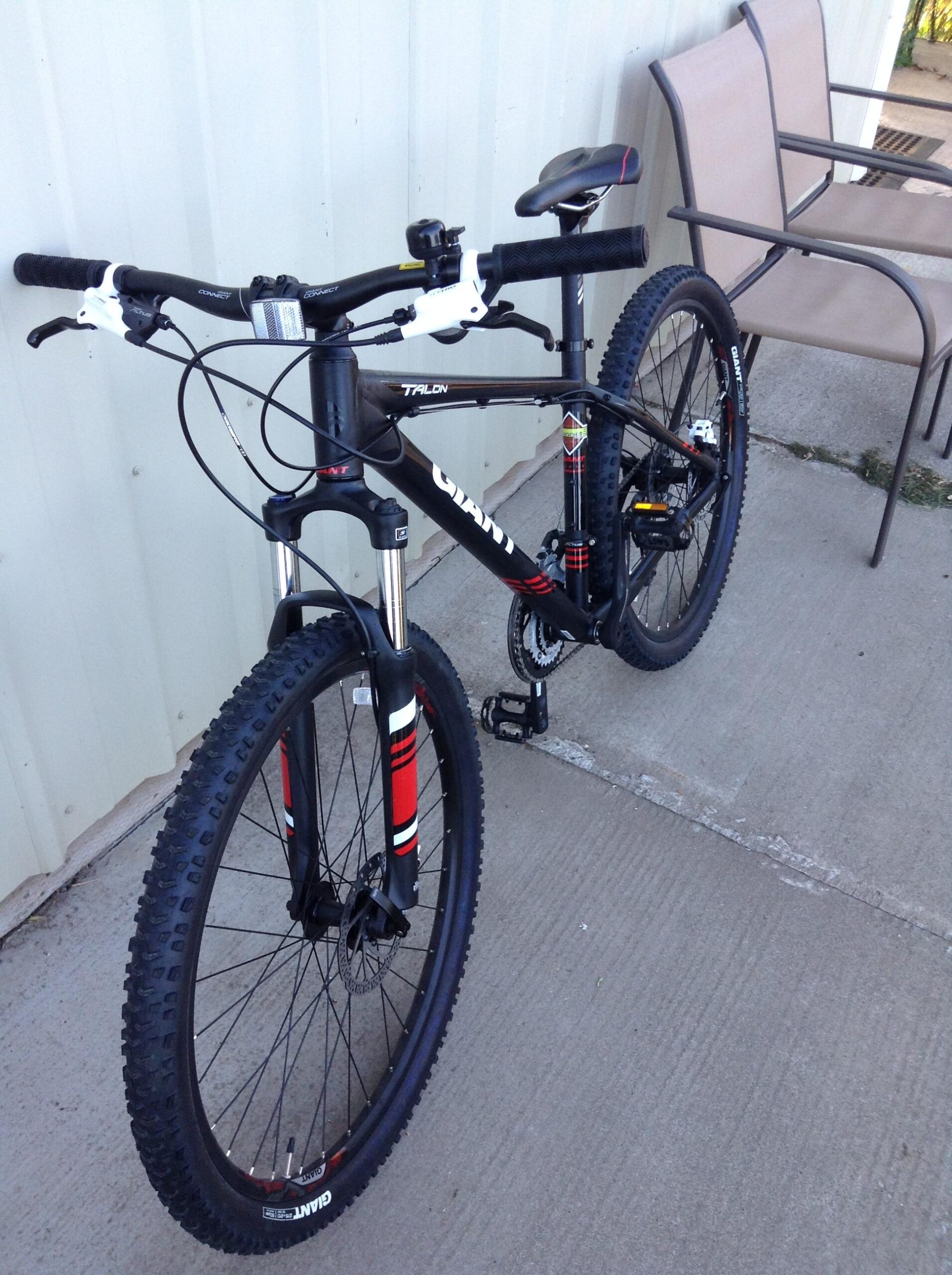 Giant Talon 27.5 3: A black and red mountain bike with wide tires, positioned on a concrete surface near a wall. The bike features a sleek frame, dual disc brakes, and prominent handlebars. In the background, there is a pair of metal chairs partially visible.