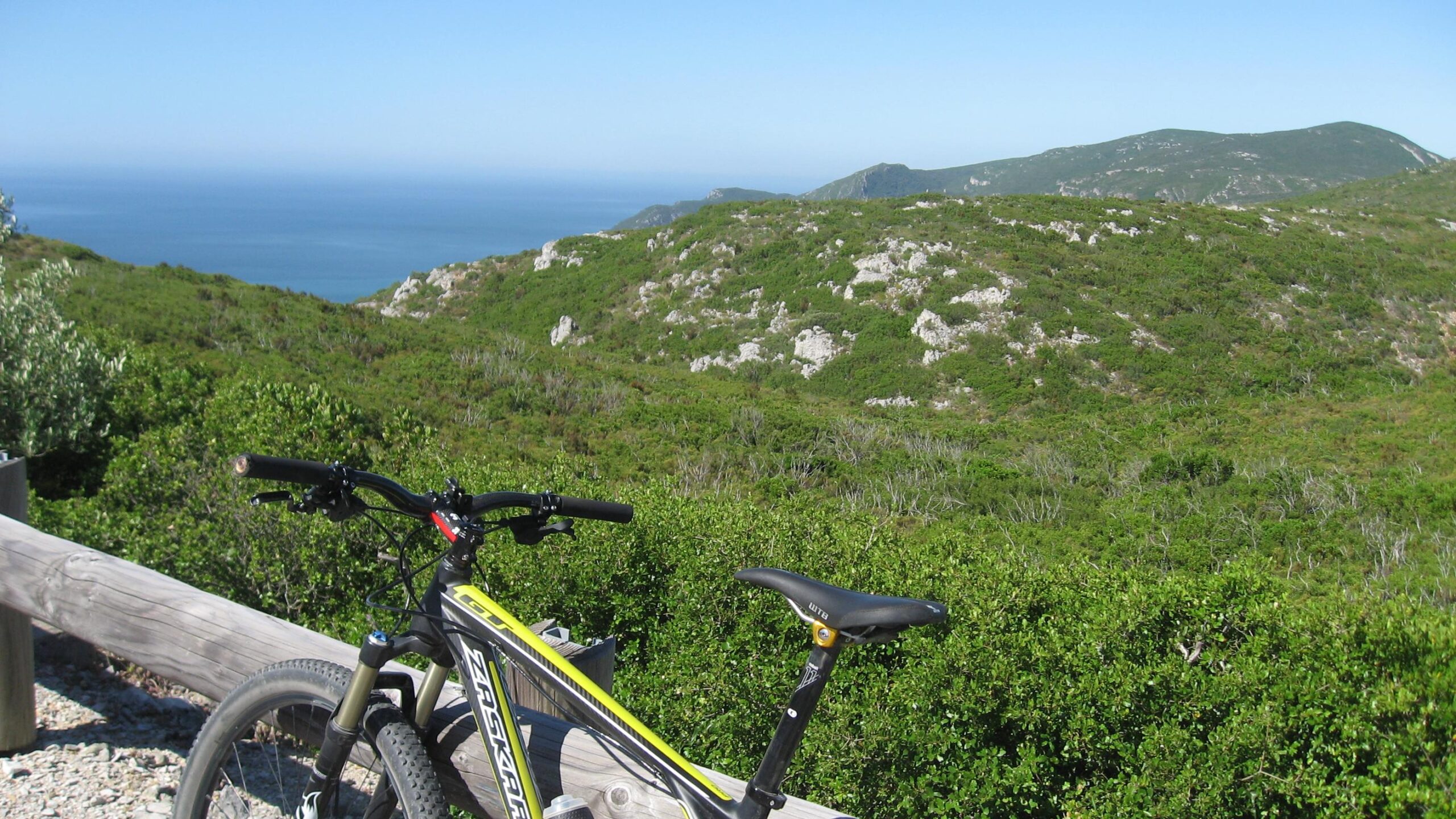 A mountain bike resting on a wooden fence overlooking a lush green landscape that slopes down to a blue ocean under a clear sky. Arrabida mountain bike trail.