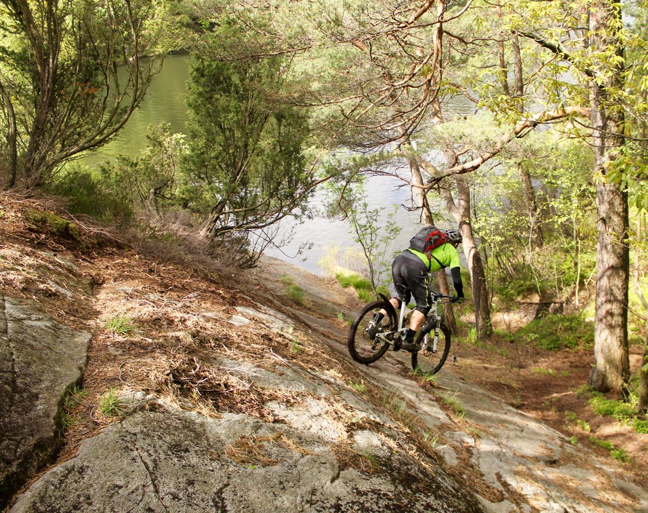 A mountain biker navigating a rocky slope near a serene lake, surrounded by trees and greenery. The rider is wearing a bright green shirt and has a backpack, showcasing an adventurous outdoor experience. Skatas Trail System mountain bike trail.