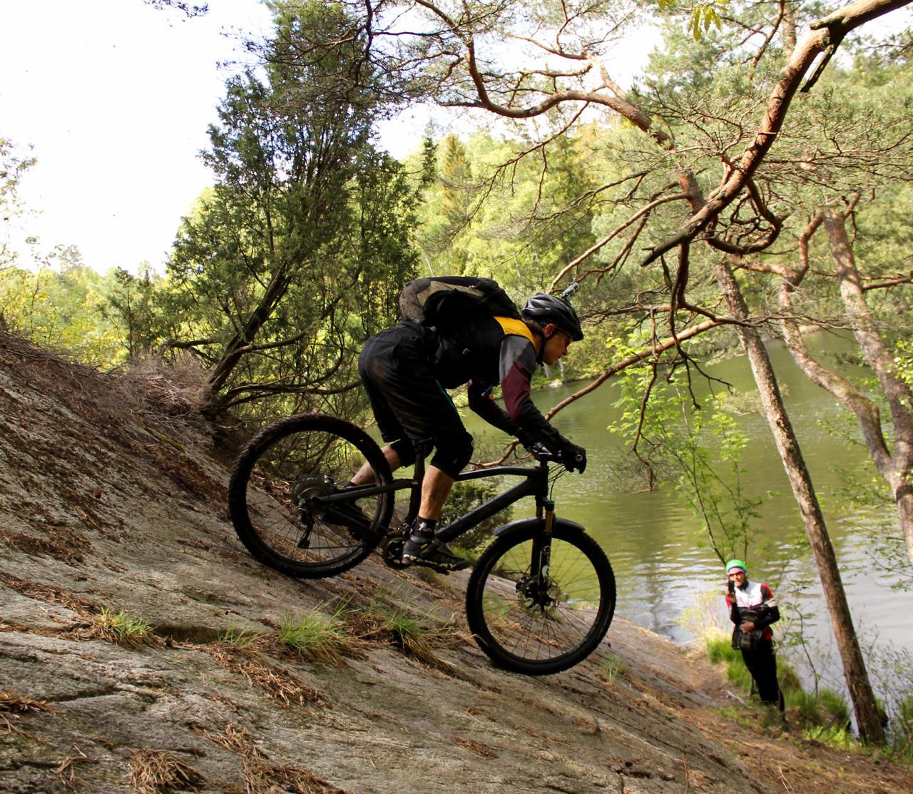 A mountain biker descends a steep, rocky slope beside a serene lake, with lush green trees in the background. Another person in cycling gear observes from a lower vantage point, creating a sense of adventure in a natural setting. Skatas Trail System mountain bike trail.