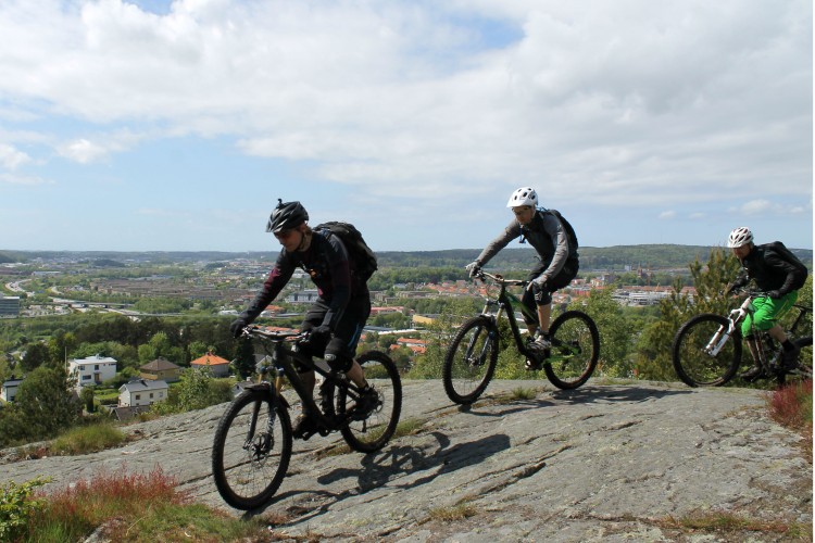 Three mountain bikers navigate rocky terrain on a hillside, with a scenic view of a town and lush greenery in the background. The cyclists are wearing helmets and protective gear, showcasing an active outdoor adventure on a bright day.