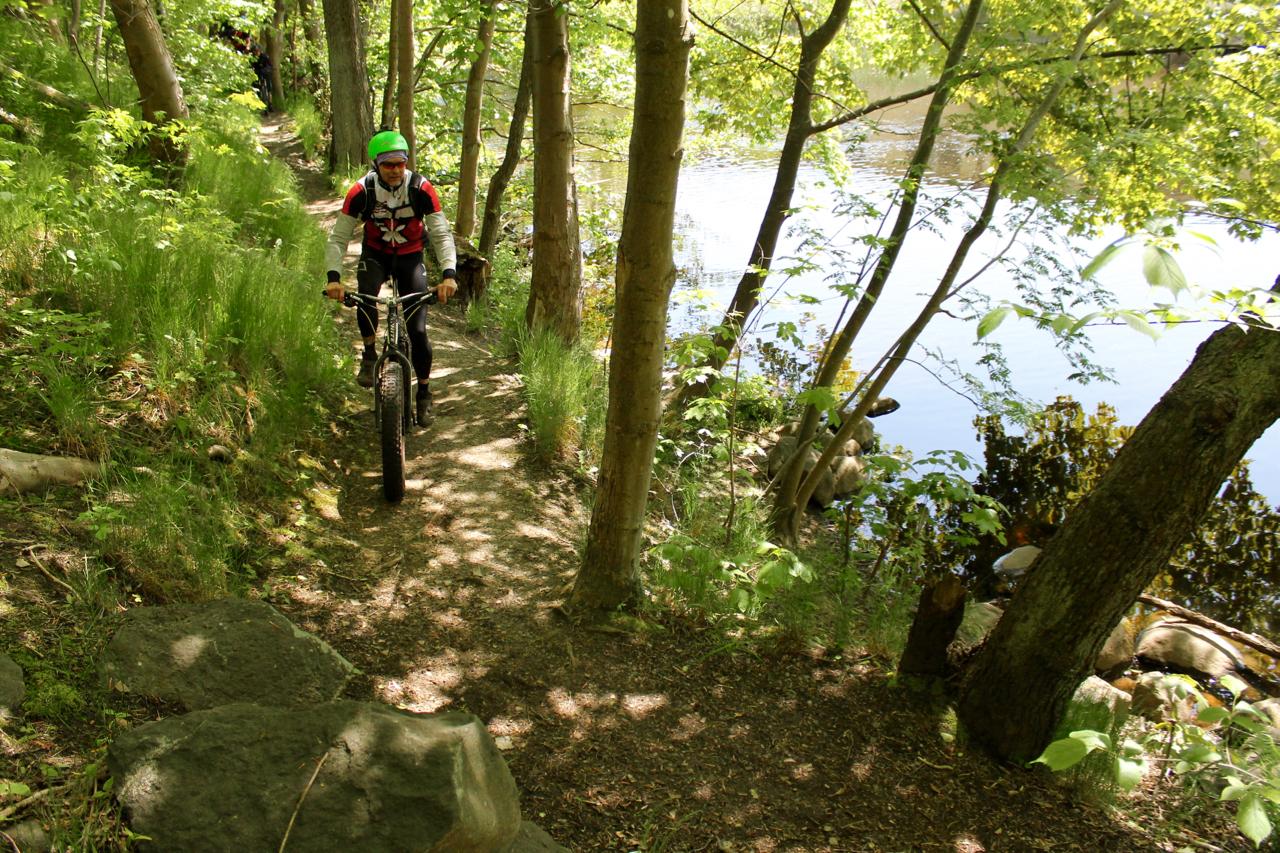 A person riding a mountain bike along a narrow, tree-lined path next to a calm body of water, surrounded by vibrant green foliage and patches of sunlight filtering through the trees. Molndal Area mountain bike trail.