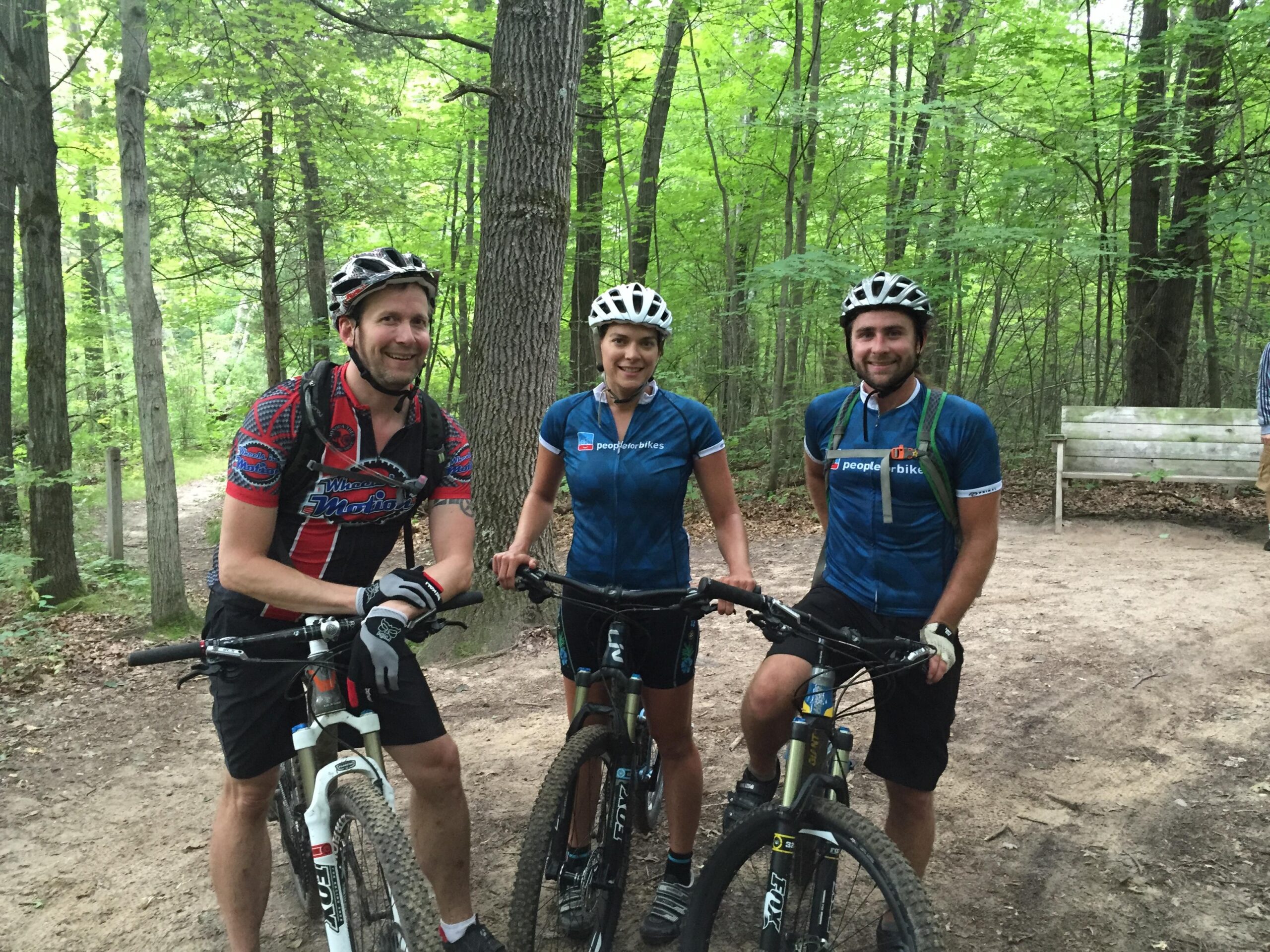 Three mountain bikers posing together on a forest trail, surrounded by green trees. They are wearing cycling jerseys and helmets, smiling and holding their bikes. A wooden bench is visible in the background along the trail. Potawatomi trail mountain bike trail.