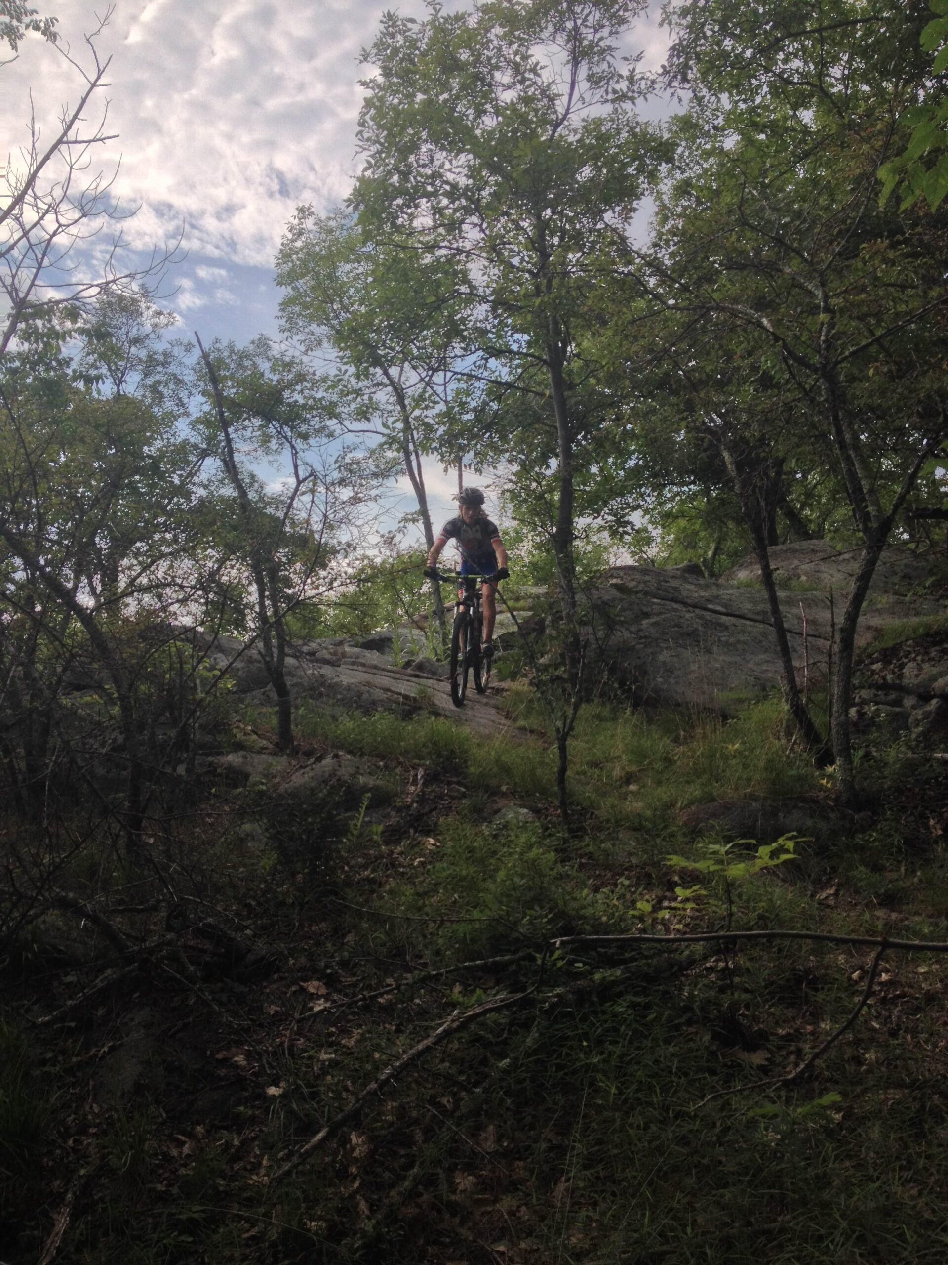 A mountain biker navigating a rocky trail surrounded by greenery and trees under a cloudy sky. Allamuchy State Park-North mountain bike trail.