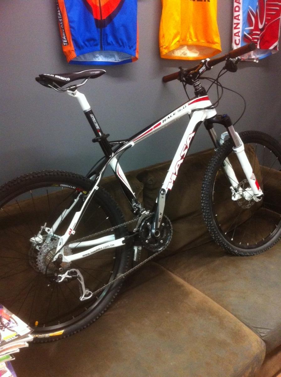 A white and black mountain bike resting on a brown couch, with its front wheel turned slightly towards the viewer. The bike is equipped with a front suspension fork and has various components visible, including the drivetrain and rear gears. In the background, brightly colored cycling jerseys are hanging on the wall.