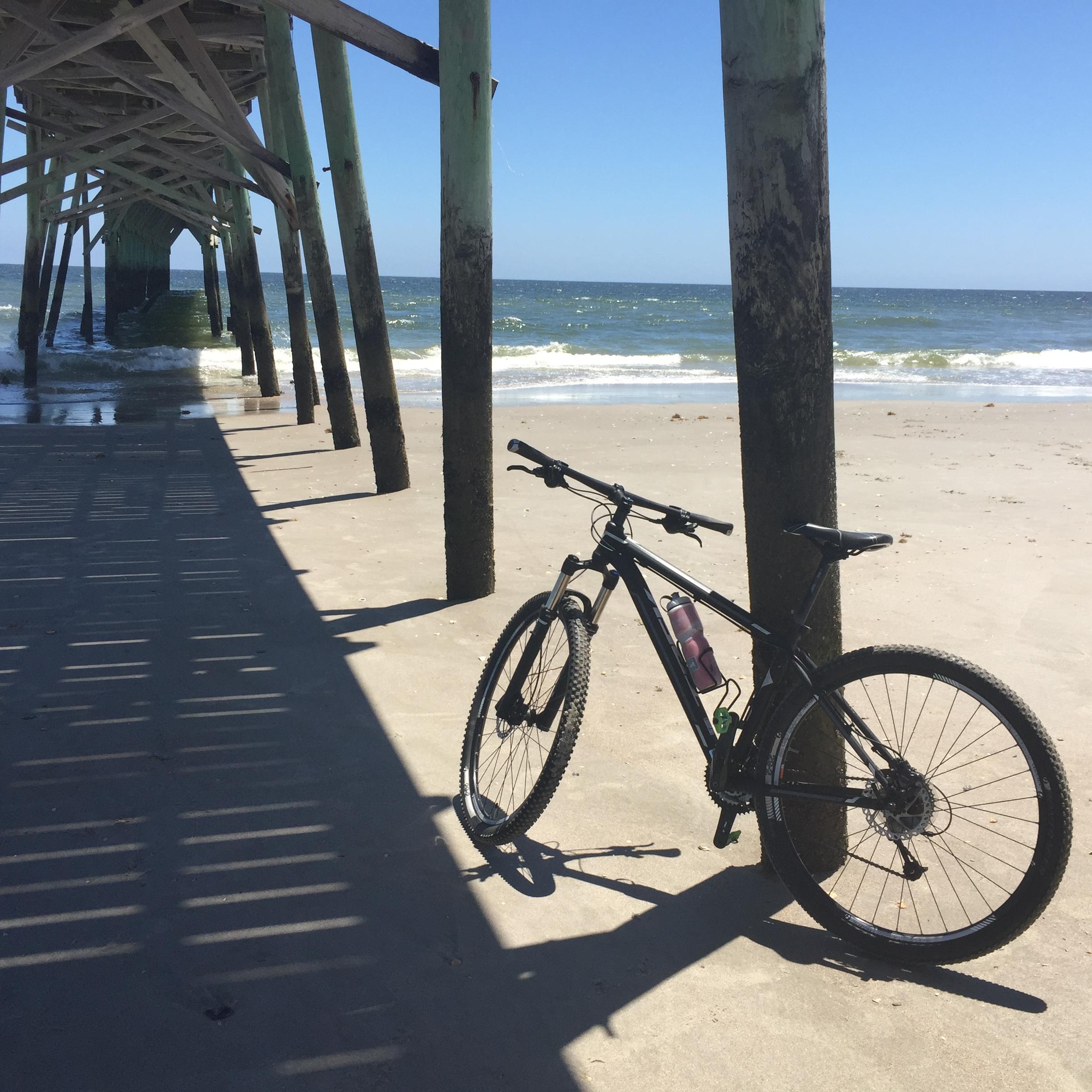 Trek Mamba Gary Fisher: A black mountain bike leaning against a wooden support pillar under a pier, with waves visible in the background and clear blue skies above. The sandy beach is partially shaded by the pier, creating a play of light and shadow on the ground.
