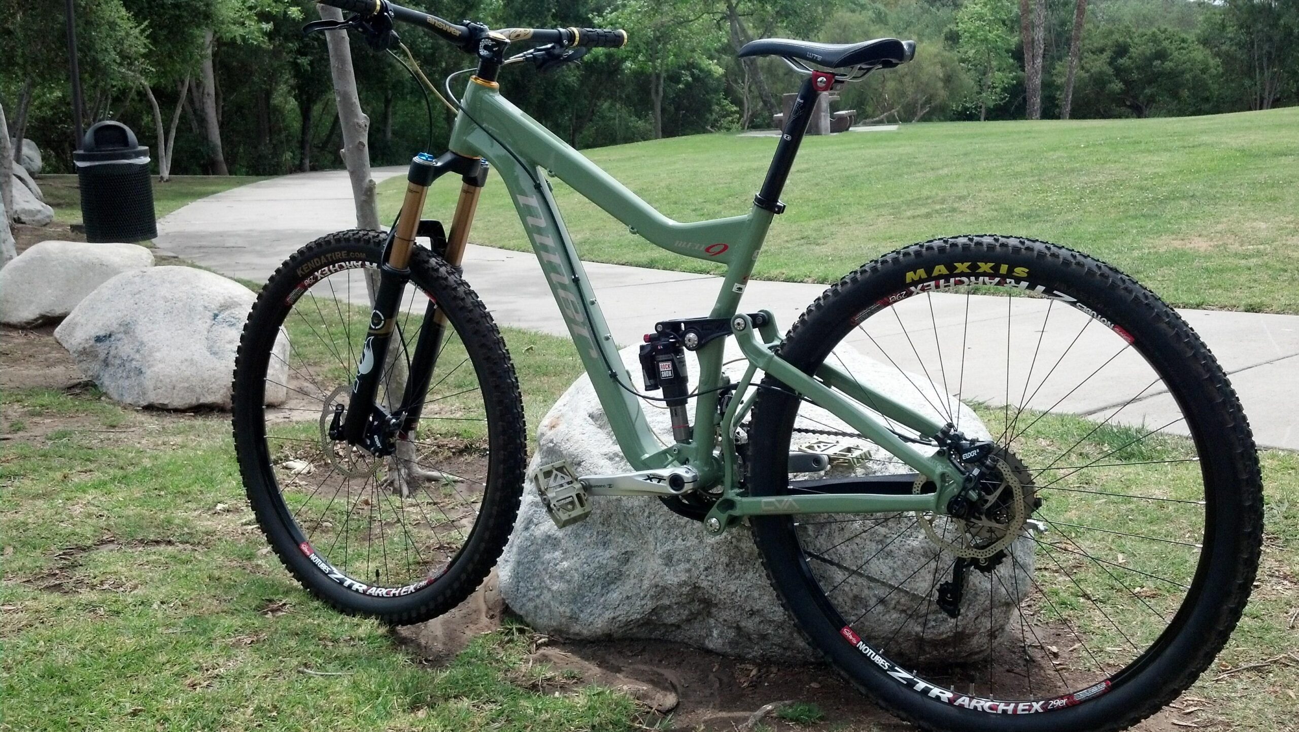 Niner W.F.O. 9: A mountain bike leaning against a large rock in a grassy area. The bike features a light green frame, black tires with prominent tread patterns, and a front suspension fork. In the background, there is a paved pathway and a trash can amidst trees and grass.